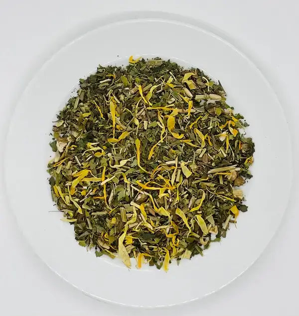 A close-up shot of loose-leaf Jaroma Goddess Bliss Tea, showcasing a blend of green tea leaves mixed with vibrant yellow calendula petals and other aromatic herbs. The tea blend exudes a calming, vibrant look on a white plate.
