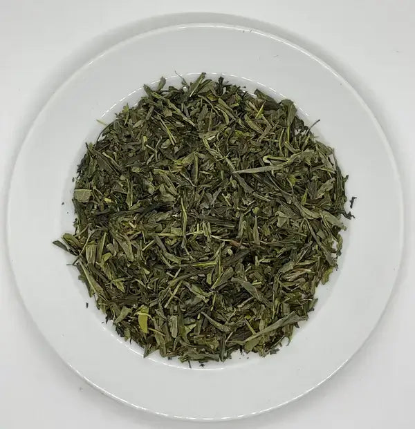 A close-up shot of dry, organic Sencha Green Tea leaves arranged on a white plate. The vibrant green leaves have a delicate, fresh appearance, capturing the essence of high-quality green tea with its earthy, refreshing fragrance