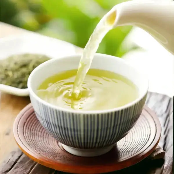A hand pours hot water into a bowl of Sencha Green Tea, creating a vibrant green infusion. The tea is steeping in a traditional blue-striped Japanese teacup placed on a wooden saucer, surrounded by loose tea leaves in the background. The warm, calming atmosphere emphasizes the fresh, organic quality of the tea