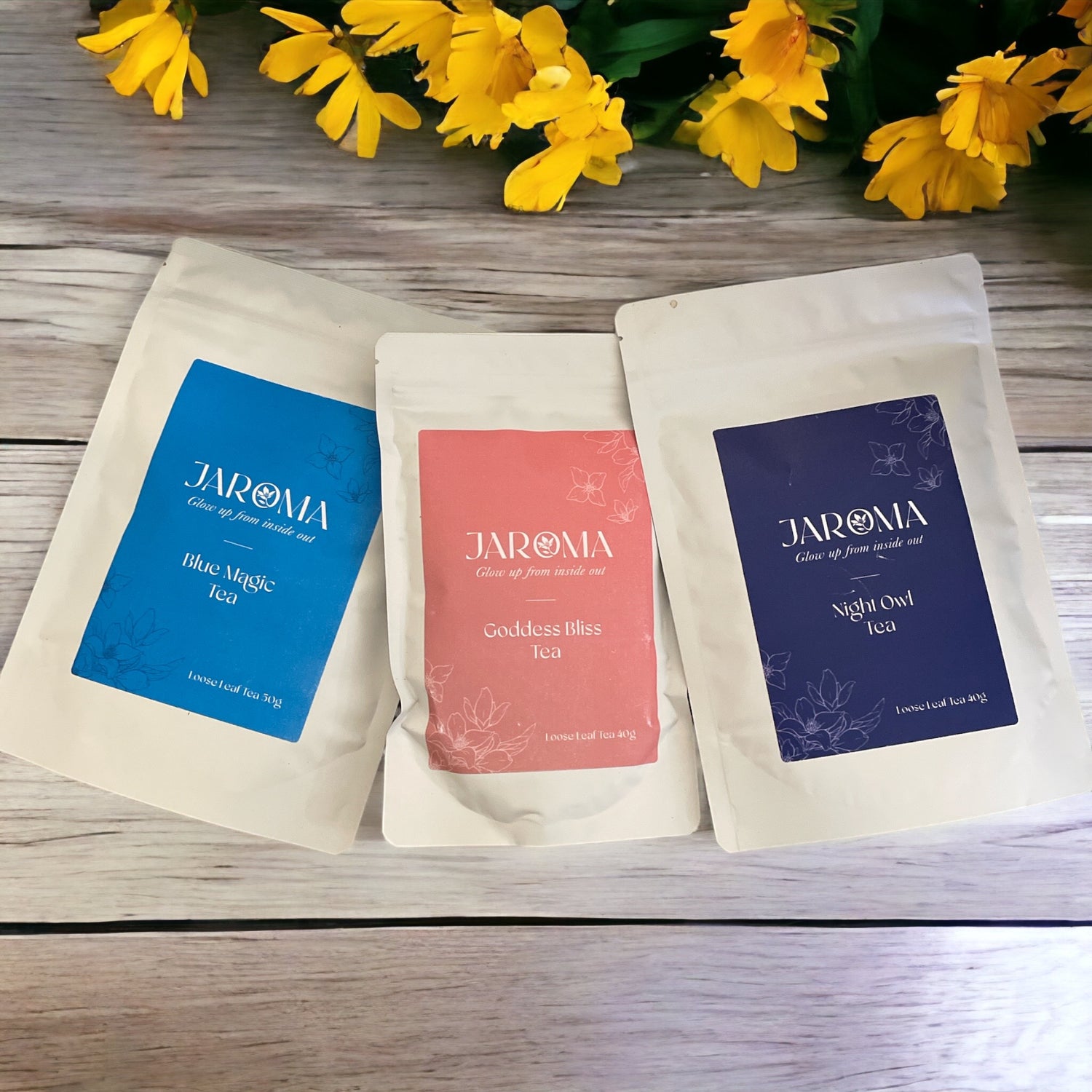 Three tea pouches from Jaroma Tea: Blue Magic, Goddess Bliss, and Night Owl, with yellow flowers in the background.